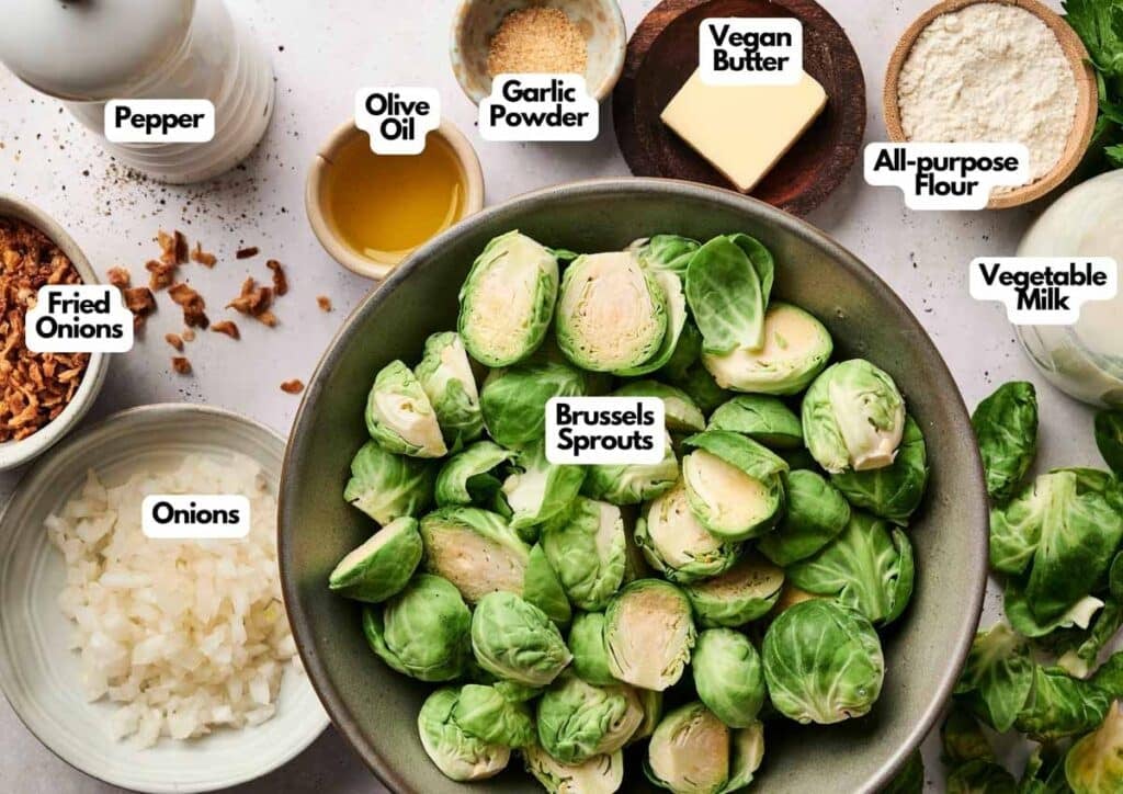 A bowl of halved Brussels sprouts for a delicious Brussels Sprout Casserole, surrounded by labeled ingredients: fried onions, chopped onions, pepper, olive oil, garlic powder, vegan butter, all-purpose flour, and vegetable milk.