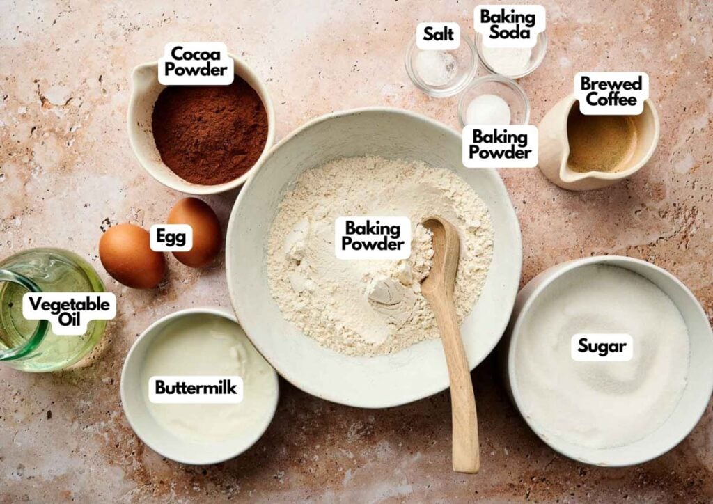 A top-down view of baking ingredients in bowls and cups for a Chocolate Cake Recipe, including flour, cocoa powder, sugar, buttermilk, eggs, vegetable oil, salt, baking powder, baking soda, and brewed coffee.