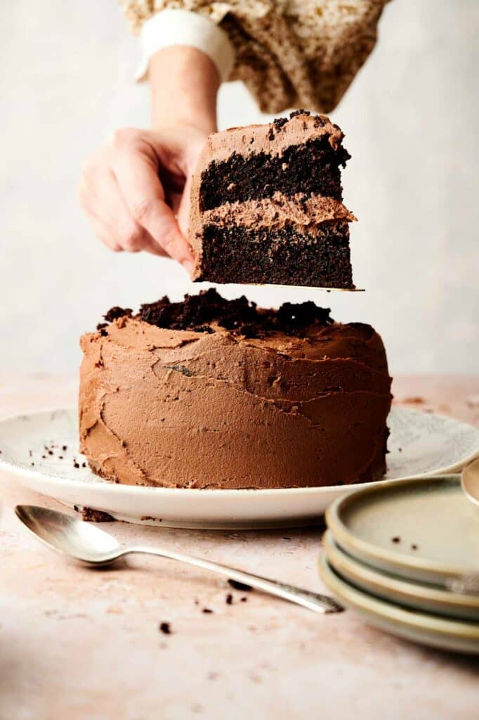 A person serving a slice of chocolate cake from a layered chocolate cake recipe on a plate.