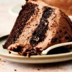 A slice of chocolate cake from a chocolate cake recipe with frosting on a plate with a fork taking a piece.