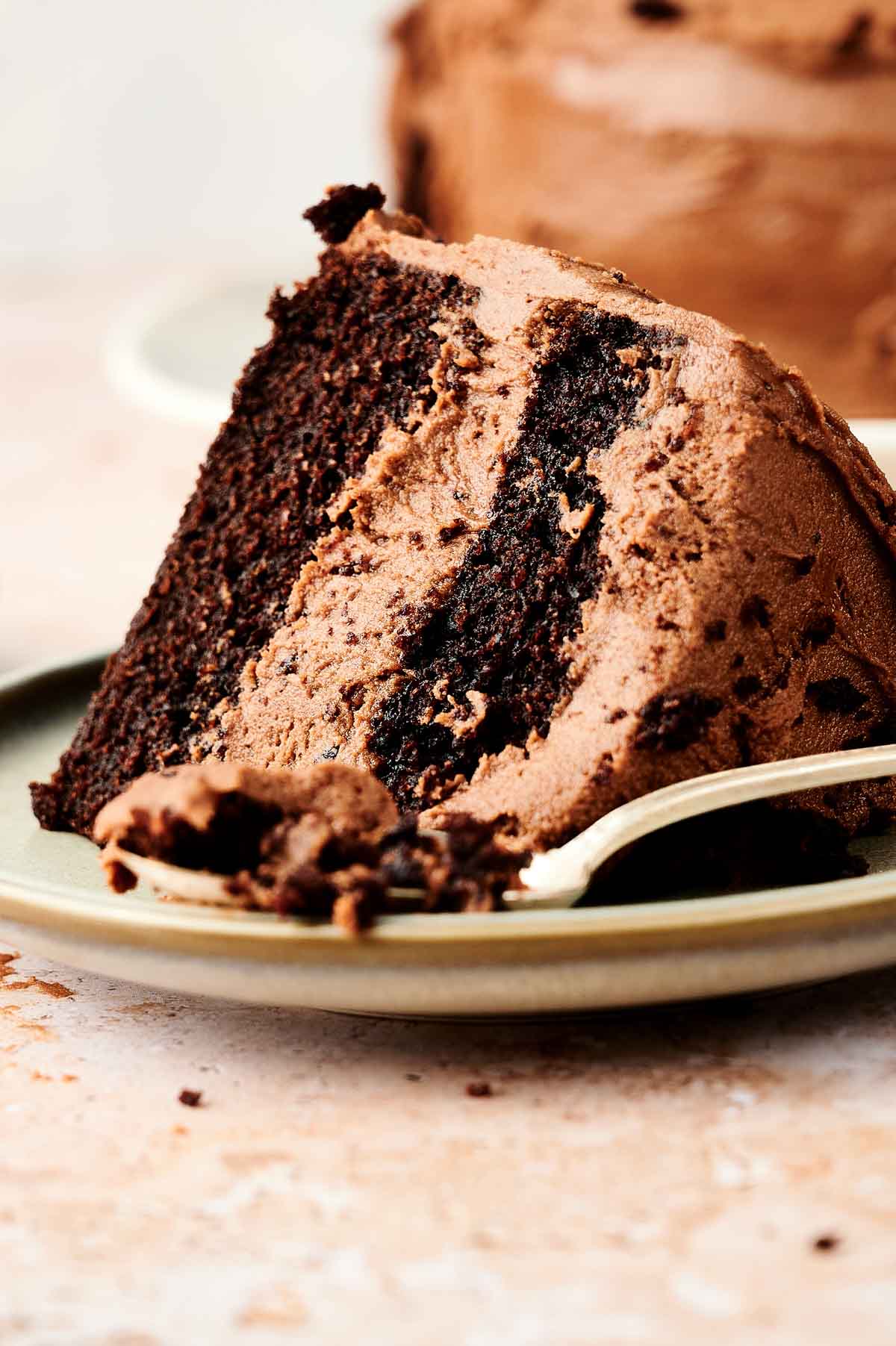 A slice of chocolate cake from a chocolate cake recipe with frosting on a plate with a fork taking a piece.