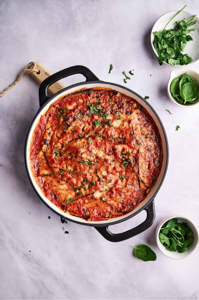 A pan of baked eggplant parmesan, reminiscent of a classic eggplant casserole, is topped with tomato sauce and herbs and surrounded by small bowls of fresh greens on a light surface.