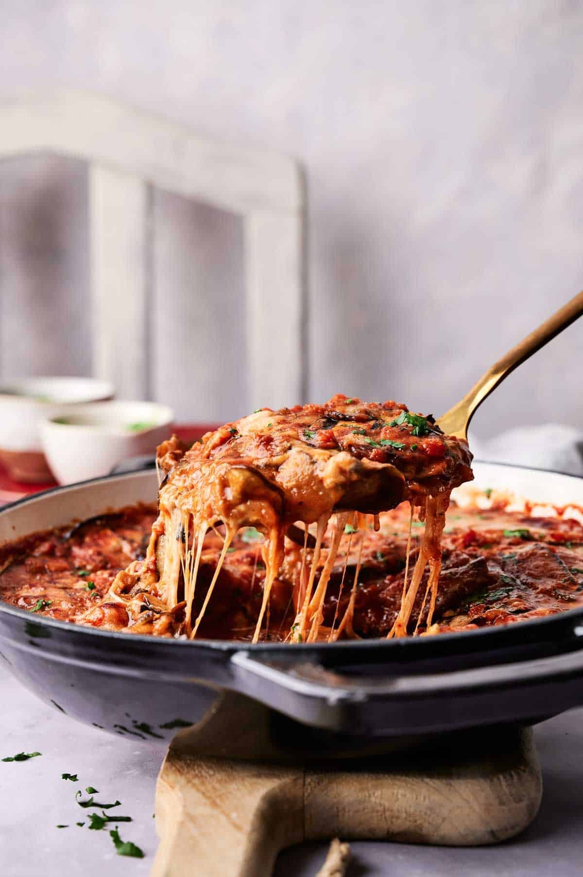 A serving of cheesy baked Eggplant Casserole is lifted from a casserole dish with a spatula, showing melted cheese stretching from the pan.
