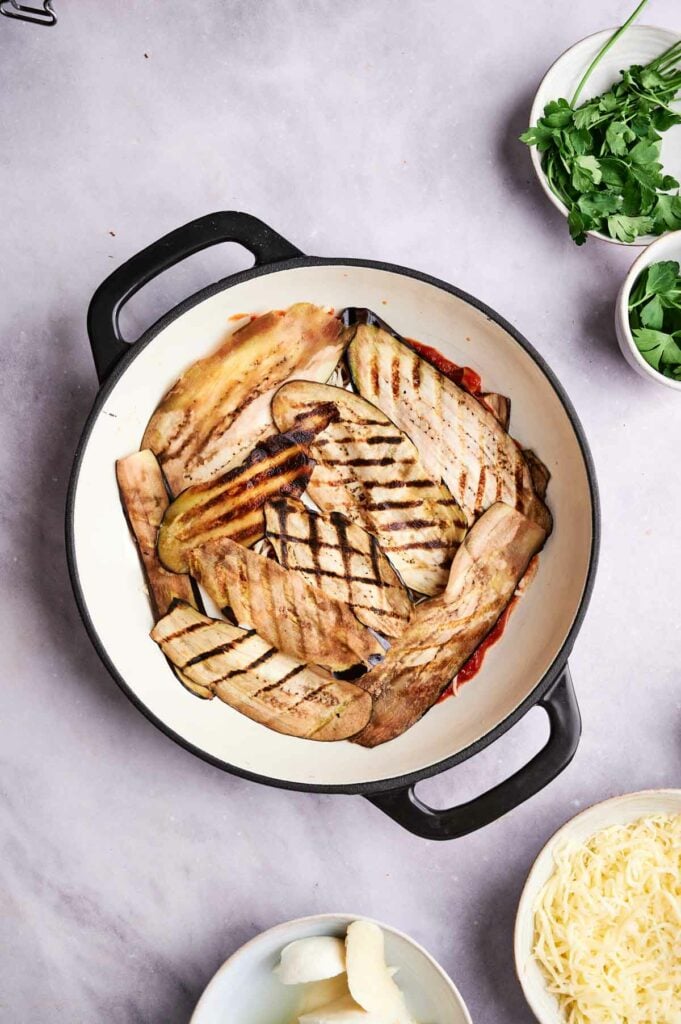 A pan of eggplant casserole with a couple bowls of parsley and a bowl of cheese.