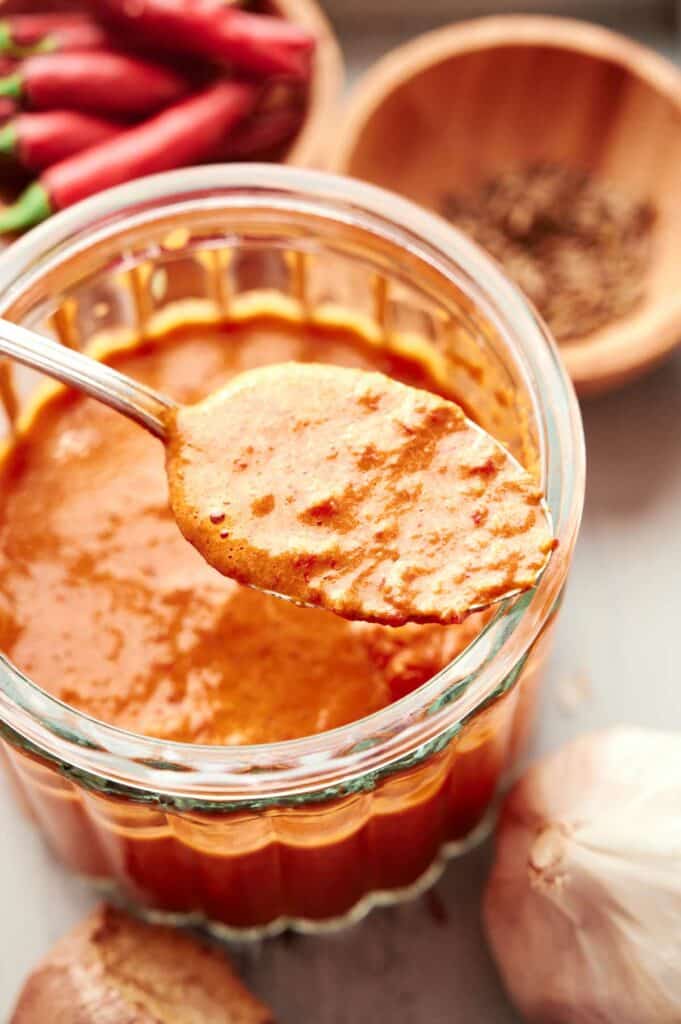 A spoon lifting Thai red curry paste from a glass jar, with garlic, chilies, and spices in the background.