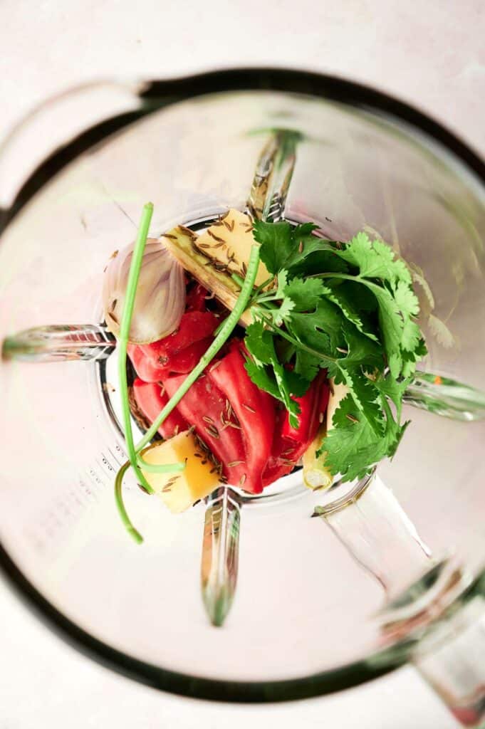 Top-down view of a blender containing fresh ingredients like tomatoes, herbs, and possibly fruits or vegetables for making a Thai Red Curry sauce.