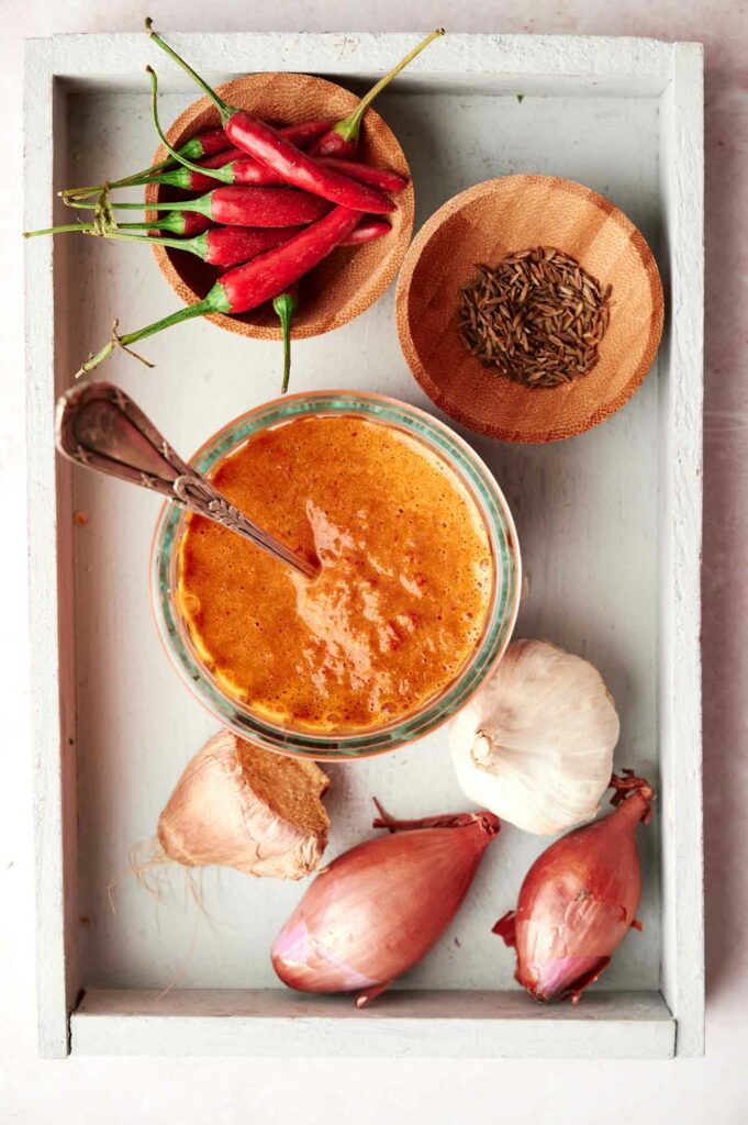 Assorted ingredients for a recipe including a jar of sauce, red chili peppers, garlic, ginger, shallots, and spices on a wooden tray.