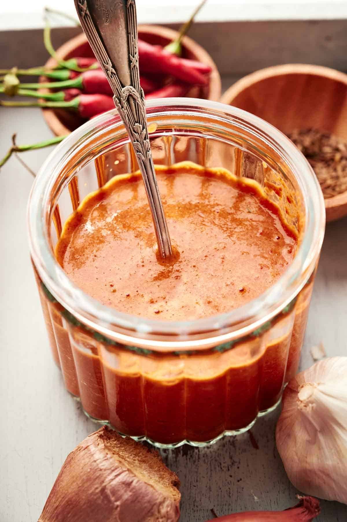 Thai red curry paste in a glass jar with a spoon, surrounded by fresh ingredients.