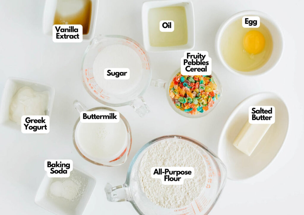 Various baking ingredients, including vanilla extract, sugar, oil, egg, Greek yogurt, buttermilk, Fruity Pebbles cereal for a Fruity Pebbles Donut, salted butter, baking soda, and all-purpose flour, are labeled on a white surface.