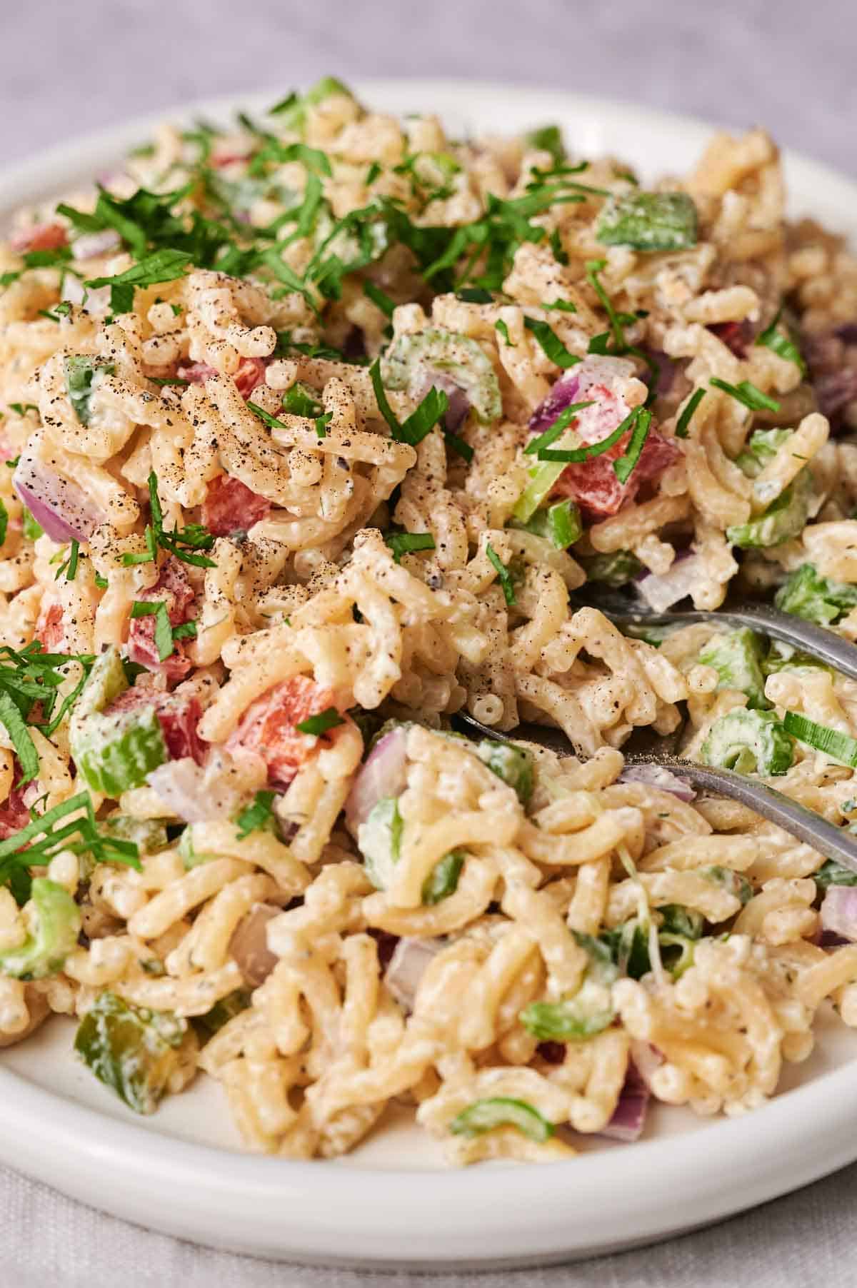 A plate of creamy macaroni salad with diced tomatoes, chopped parsley, and onions, seasoned with black pepper, served with a fork.