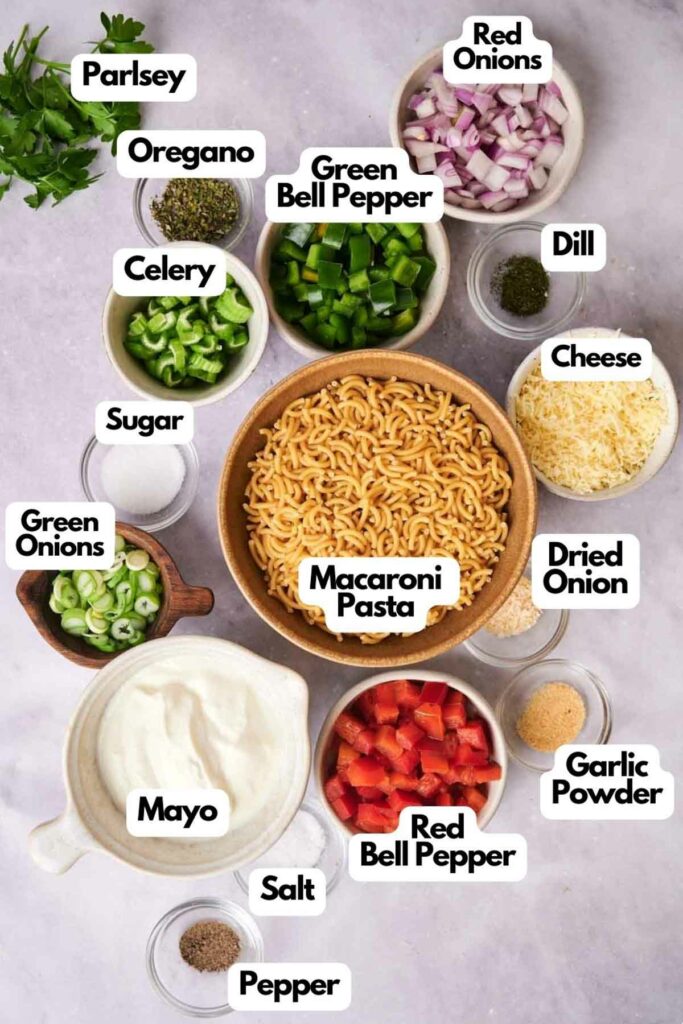 Various ingredients for pasta salad preparation, including noodles, red onions, green bell peppers, and seasonings, organized in labeled bowls on a table.