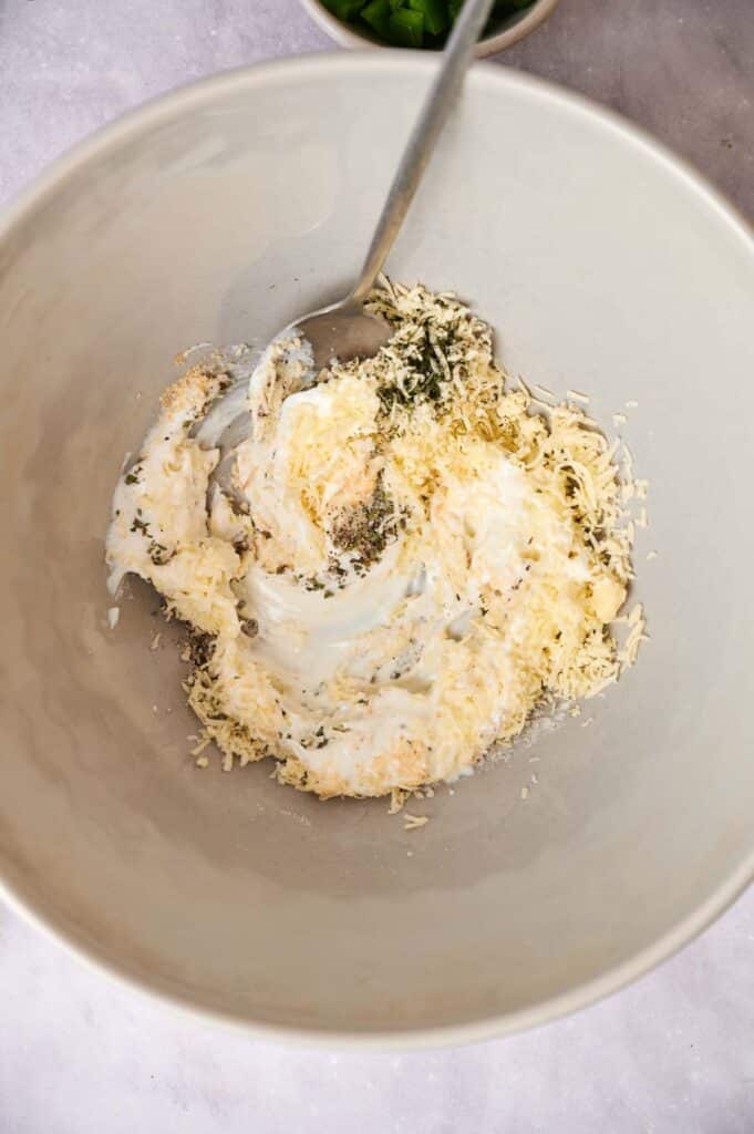 A bowl containing a mixture of cream cheese, herbs, and grated cheese with a mixing spoon.