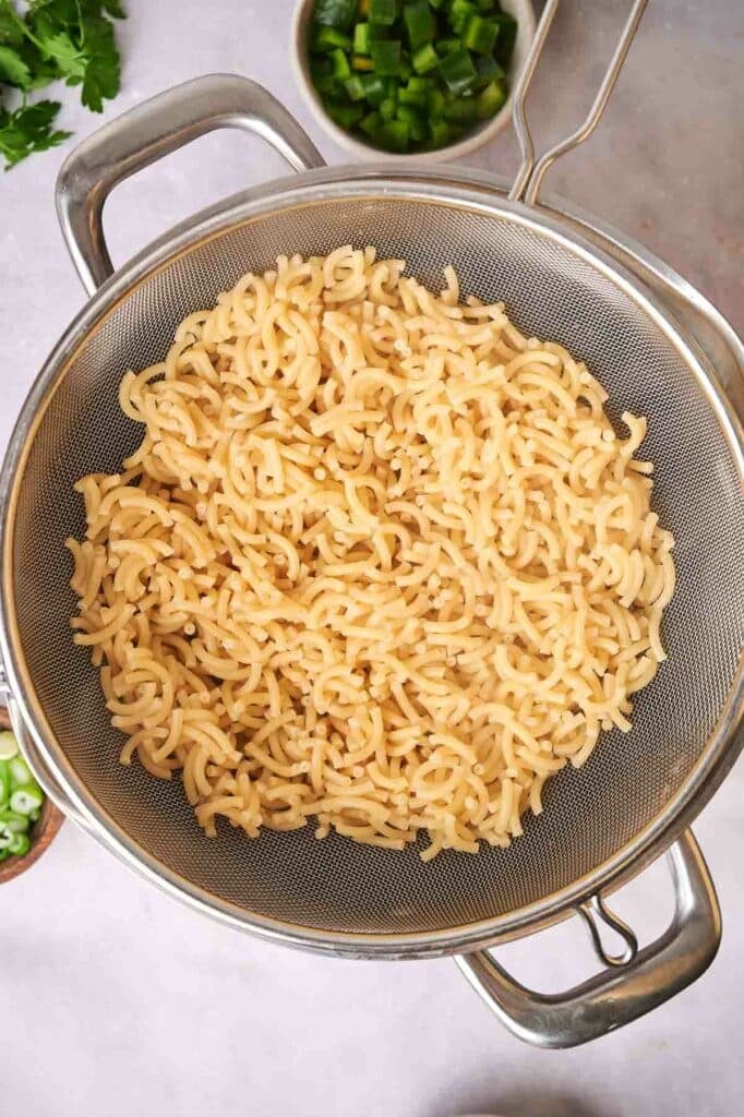 Cooked instant pasta draining in a metal strainer over a bowl, with chopped green vegetables and parsley nearby.