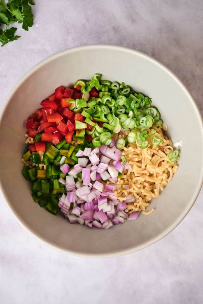 A bowl containing chopped red bell peppers, green onions, celery, red onions, and garlic on a marble surface.