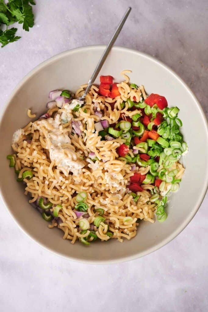 A bowl of macaroni salad with diced red peppers, green onions, and a creamy dressing, served with a fork on a light background.