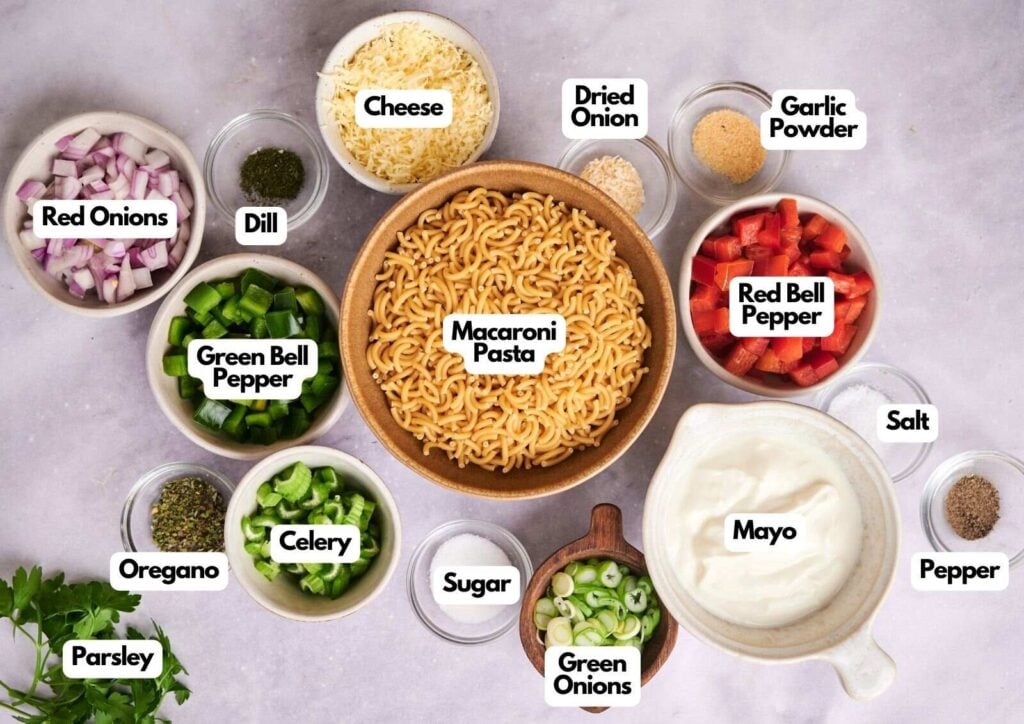 Bowls of labeled ingredients for Macaroni Salad, including pasta, vegetables, herbs, seasonings, cheese, and mayonnaise, are arranged on a light surface.