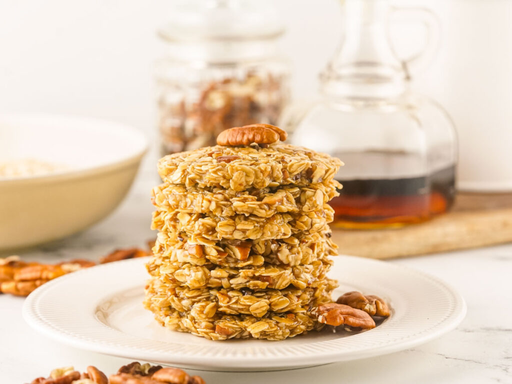 A stack of No Bake Maple Pecan Cookies on a white plate, with pecans scattered around and jars of syrup and nuts in the background.