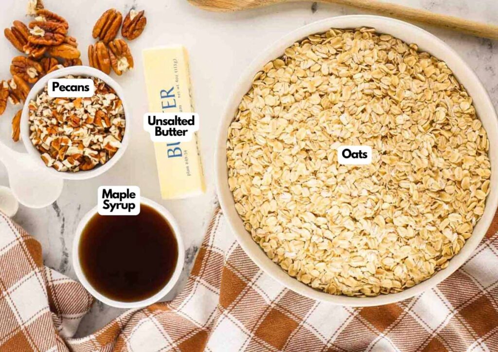 A bowl of oats, a stick of unsalted butter, a bowl of pecans, and a bowl of maple syrup on a checkered cloth with a wooden spoon—perfect ingredients for No Bake Maple Pecan Cookies.