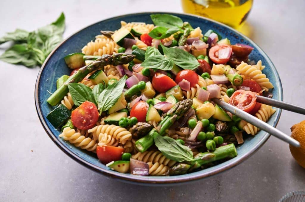 A vibrant Pasta Primavera salad with tomatoes, asparagus, peas, red onions, and basil leaves in a blue bowl, served with tongs.