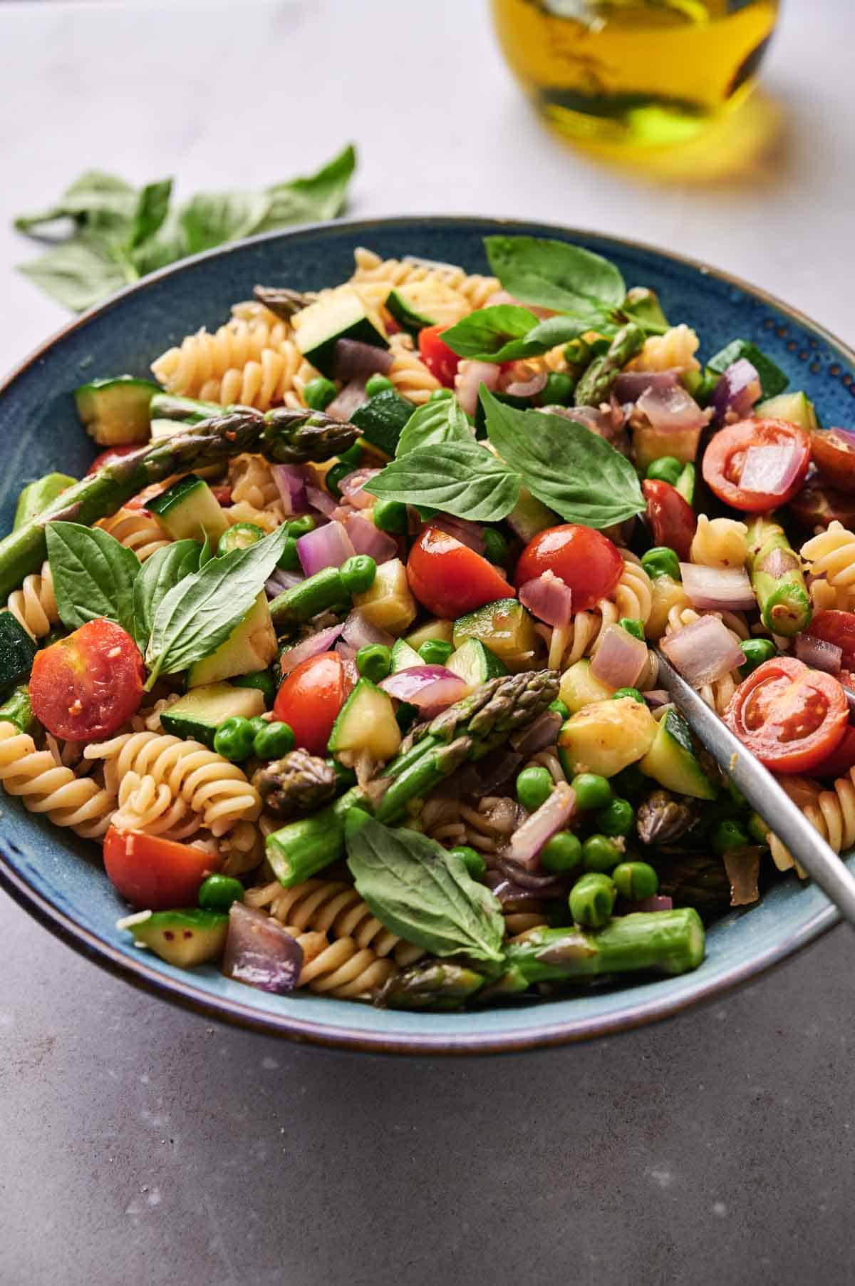 A colorful Pasta Primavera salad with fusilli, cherry tomatoes, asparagus, peas, and fresh basil, served on a blue plate with a fork.