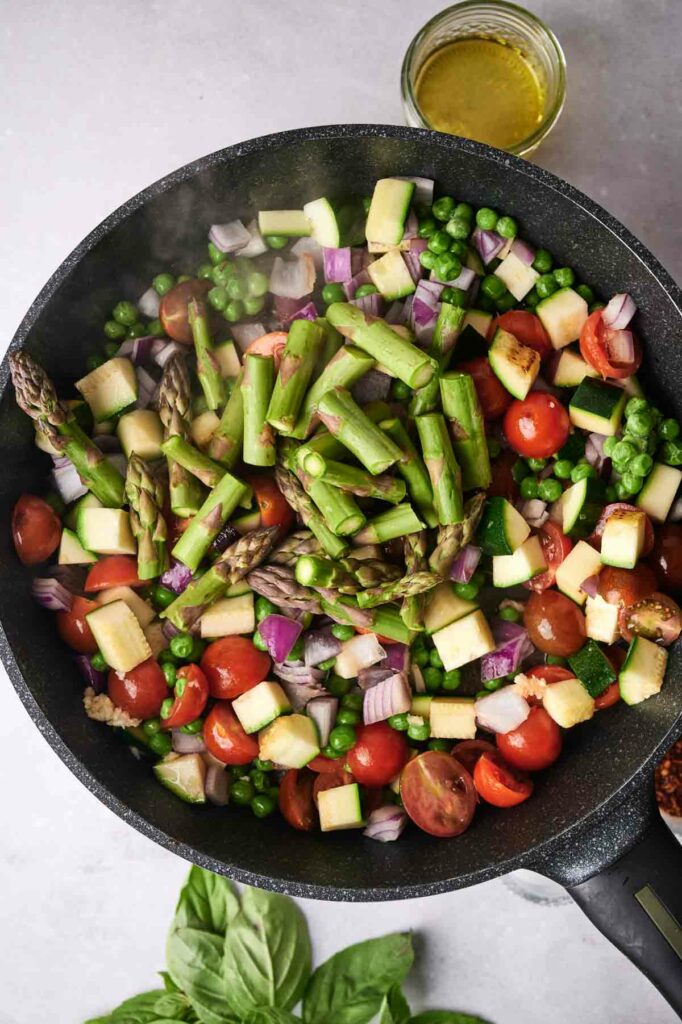 A skillet filled with colorful vegetables, including chopped asparagus, peas, tomatoes, zucchini, and red onions, on a light surface with oil and basil nearby for making Pasta Primavera.