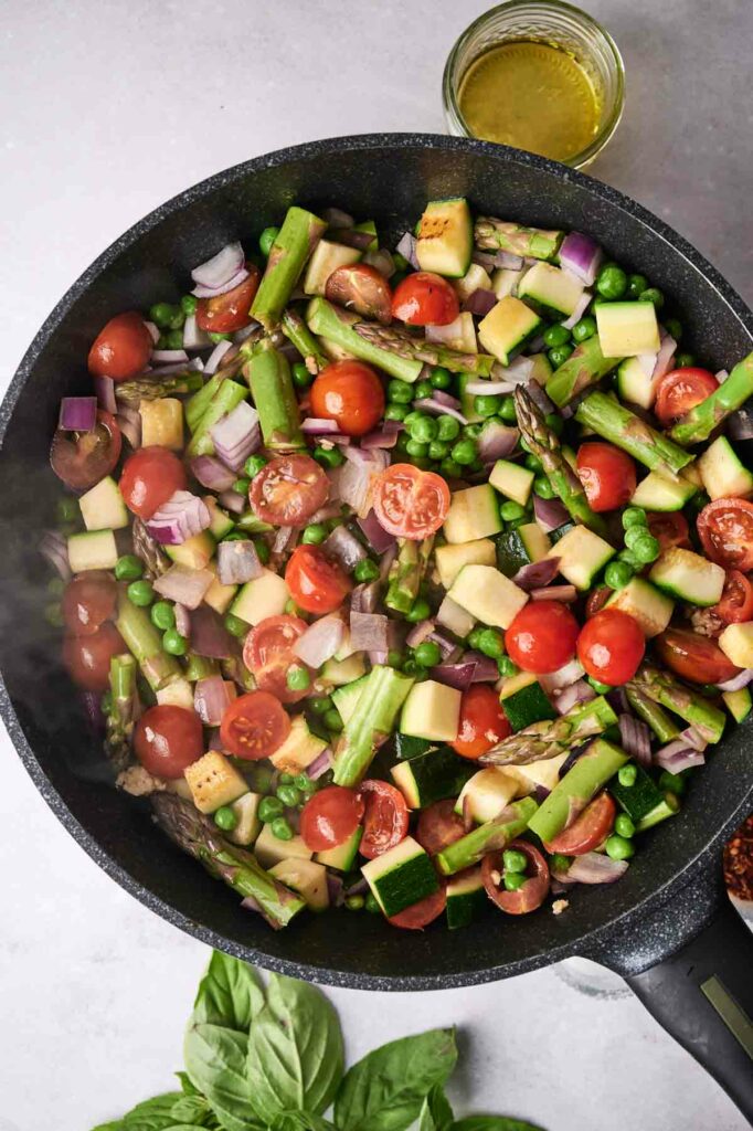 A skillet filled with sautéed vegetables including asparagus, tomatoes, onions, zucchini, and Pasta Primavera, seasoned with herbs, viewed from above.
