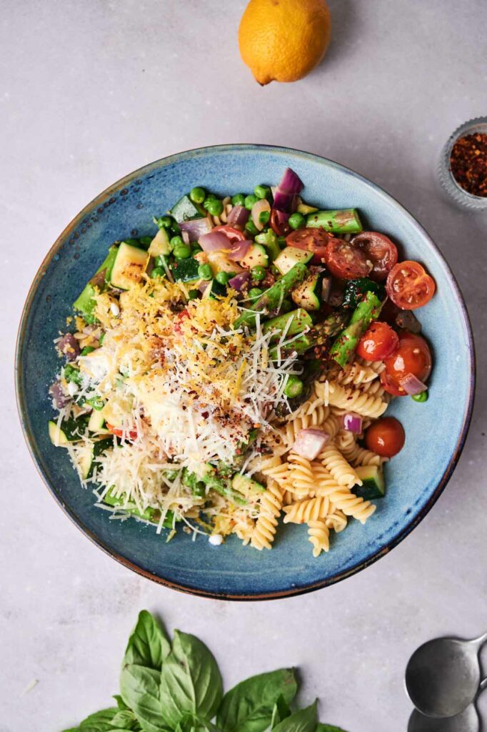 A vibrant Pasta Primavera salad with tomatoes, cucumbers, onions, and basil, topped with parmesan, served on a blue plate with a lemon half and spices in the background.