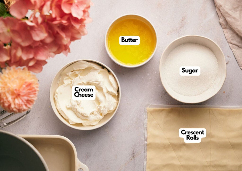 Bowls of cream cheese, melted butter, sugar, and a package of crescent rolls are arranged on a light countertop next to a pink flower bouquet.