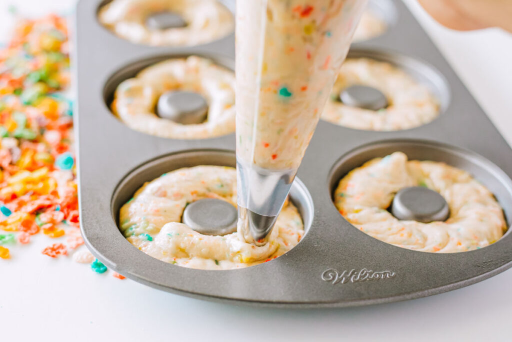 A piping bag fills a donut pan with colorful, sprinkle-filled batter for Fruity Pebbles Donut treats, with scattered cereal pieces visible on the counter.
