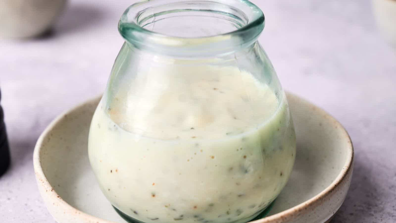 A small glass jar filled with creamy, light-colored vegan Alfredo sauce sits on a shallow ceramic dish.