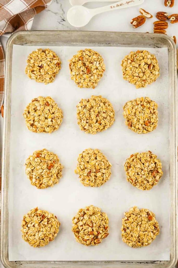 No bake maple pecan cookies with nuts and seeds on a parchment-lined tray, positioned neatly with a white spatula and scoop in the background.