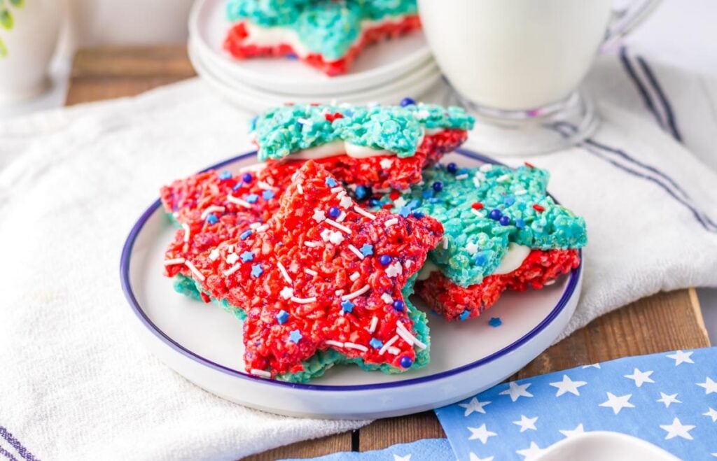 Plate with Patriotic Rice Crispy Treats in red and blue star shapes, some with white filling, topped with red, white, and blue sprinkles. A glass of milk and a star-patterned napkin sit nearby.