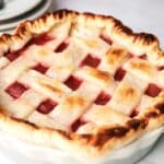 A close-up of a strawberry rhubarb pie with a lattice crust and visible red filling inside a round white dish, placed on a light-colored surface.