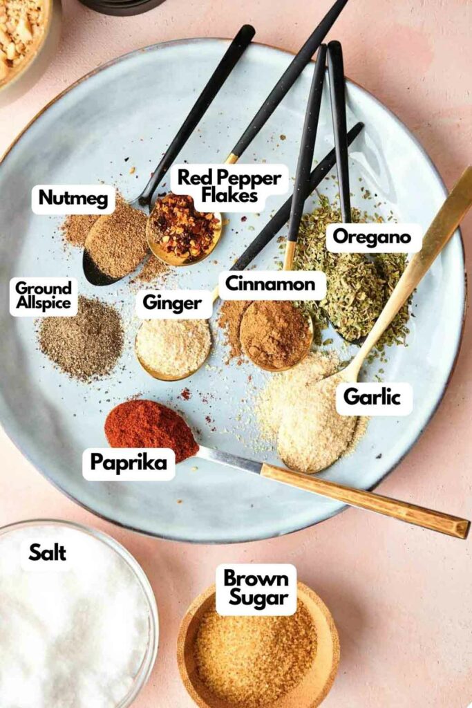 A plate with labeled spices including red pepper flakes, oregano, ground allspice, nutmeg, ginger, cinnamon, garlic, paprika, salt, and brown sugar, with spoons and a small bowl.