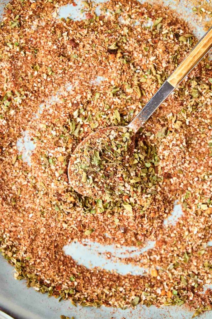 Close-up of a spoon blending a mixture of dried herbs and spices in a bowl for a jerk seasoning.
