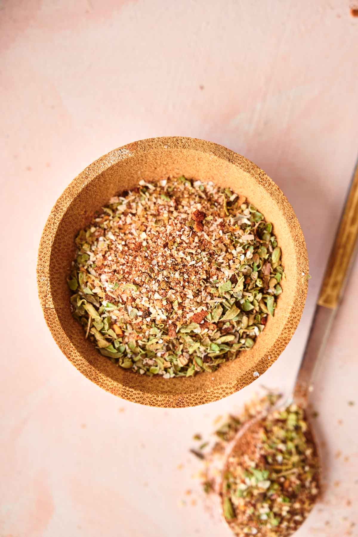 A bowl filled with a mixture of jerk seasoning and dried herbs, with a wooden spoon containing some of the mixture beside it on a light pink surface.