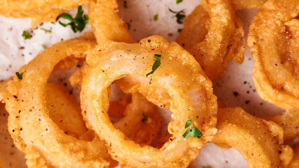 Close-up view of crispy, golden-brown onion rings seasoned with black pepper and garnished with finely chopped parsley.