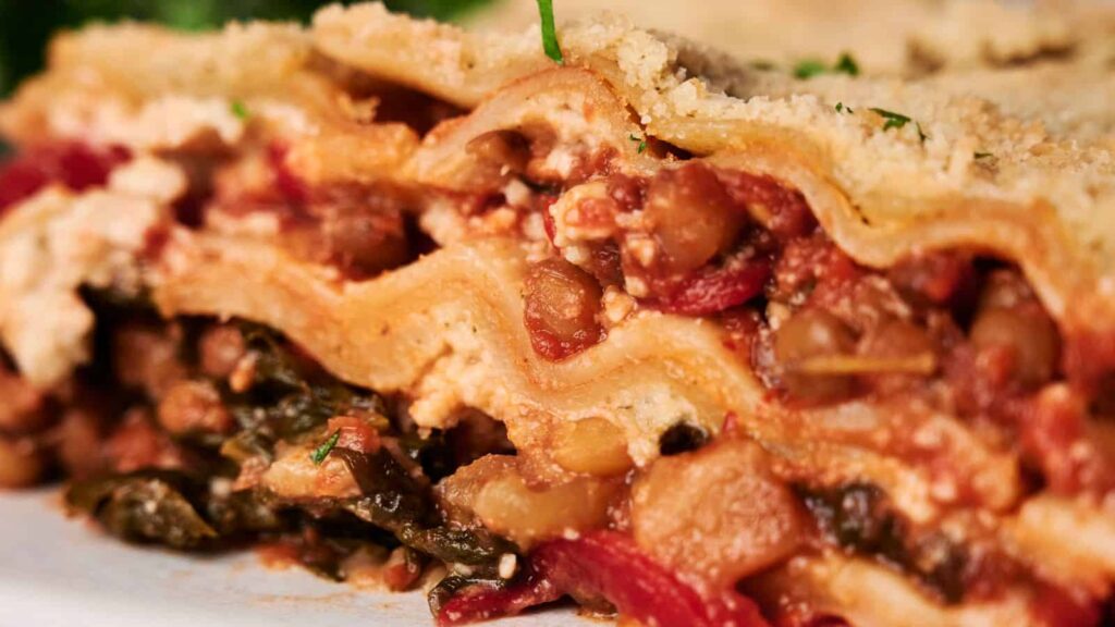 Close-up of a slice of lasagna, featuring layers of pasta, tomato sauce, vegetables, and cheese, garnished with herbs.