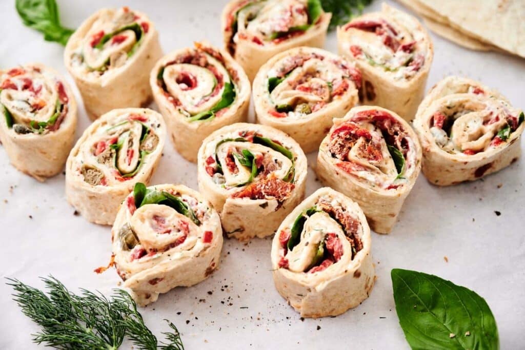 Plate of pinwheel sandwiches filled with various ingredients including greens, sliced deli meats, and cheese, arranged on a white surface with fresh herbs scattered around.