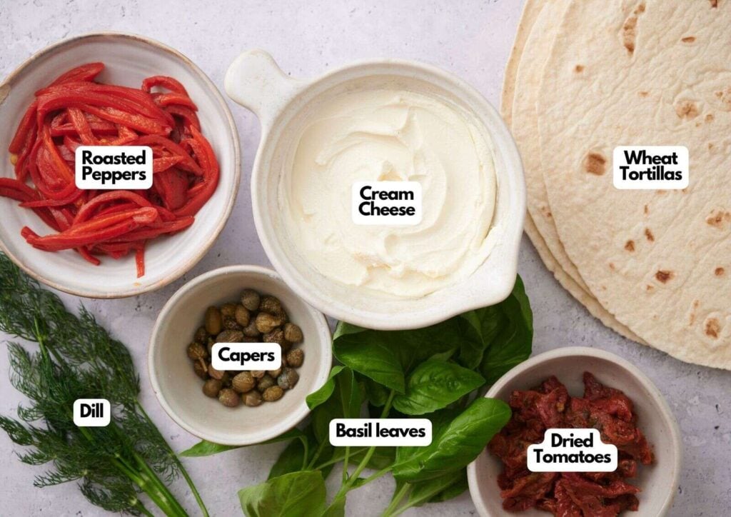 Top-down view of wheat tortillas, cream cheese, roasted peppers, capers, dried tomatoes, dill, and basil leaves arranged in bowls on a light surface—perfect ingredients for making flavorful pinwheel sandwiches.