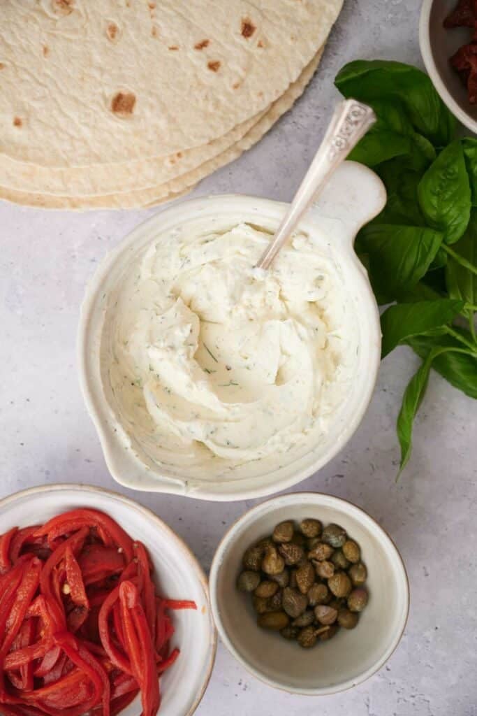 A bowl of cream cheese dip with a spoon, surrounded by red pepper strips, capers, fresh basil leaves, and a stack of tortillas.