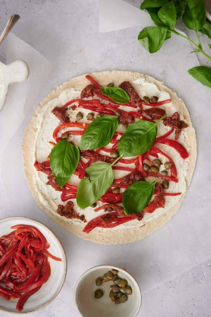 Flatbread topped with cream cheese, red bell peppers, sun-dried tomatoes, capers, and fresh basil leaves. Nearby are bowls containing sliced red bell peppers and capers.