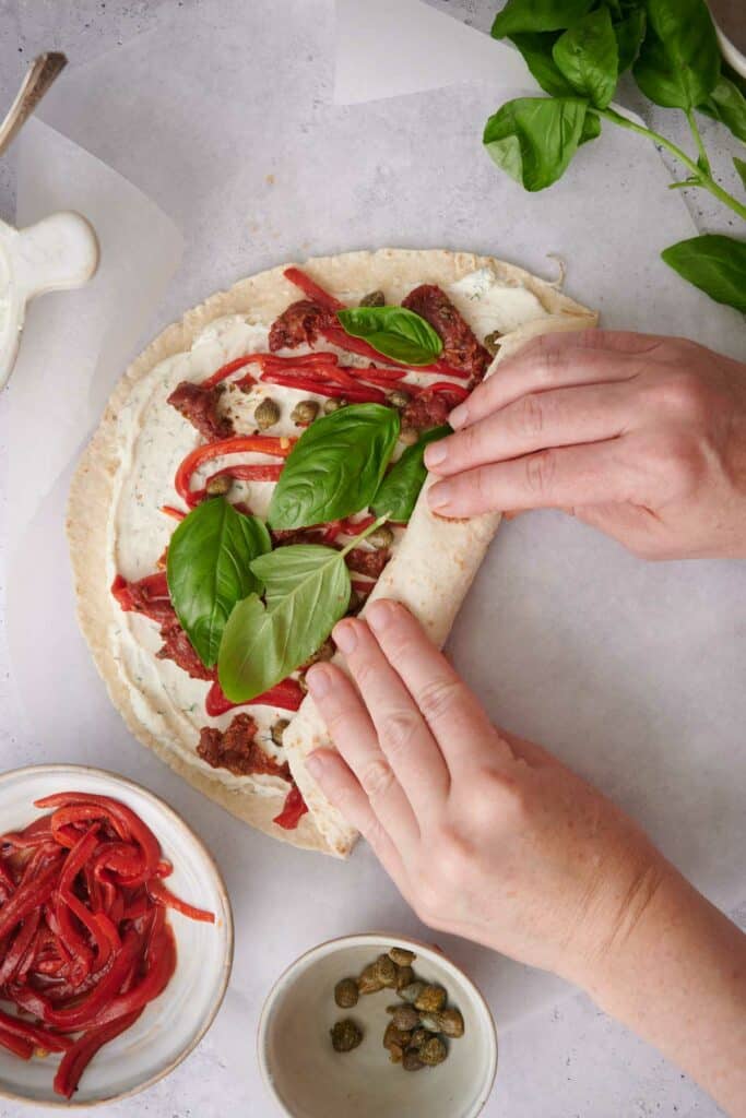 Hands rolling a tortilla wrap filled with cream cheese, sun-dried tomatoes, red bell peppers, capers, and fresh basil leaves. Bowls of ingredients are placed nearby on a light-colored surface.