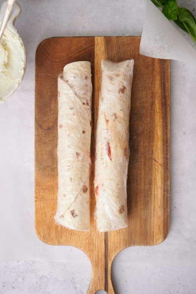 Two wrapped tortilla wraps are placed side by side on a wooden cutting board. One is slightly larger than the other, and the surface surrounding the board is white.