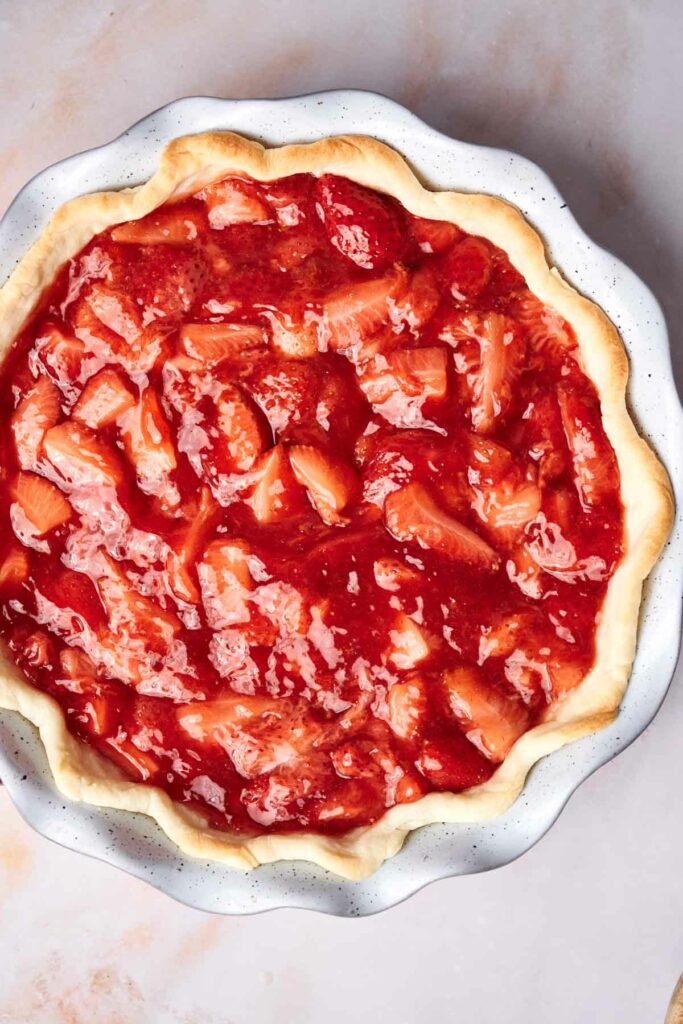 A freshly baked strawberry pie with a flaky crust, filled with bright red strawberry and rhubarb chunks, sits in a white ceramic pie dish on a light marble surface.