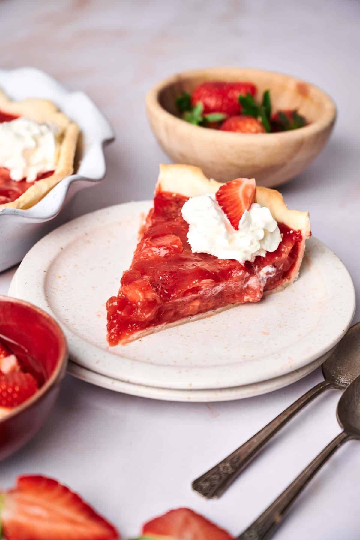 A slice of strawberry pie topped with whipped cream sits on a plate, with bowls of fresh strawberries and the whole strawberry pie in the background. Two spoons are placed beside the plate.