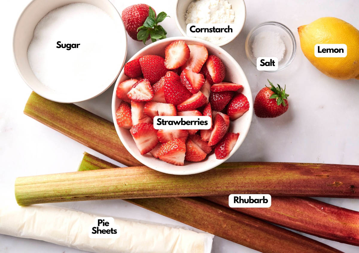 Ingredients for a classic strawberry rhubarb pie&mdash;strawberries, rhubarb, sugar, pie sheets, cornstarch, salt, and lemon&mdash;are beautifully arranged on a white surface.