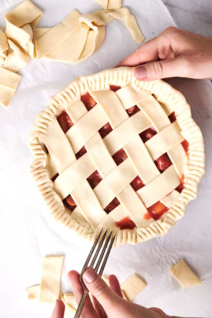 Two hands are seen preparing a lattice-topped strawberry rhubarb pie, crimping the pie's edges with a fork. Strips of excess dough are visible around the pie on a white surface.