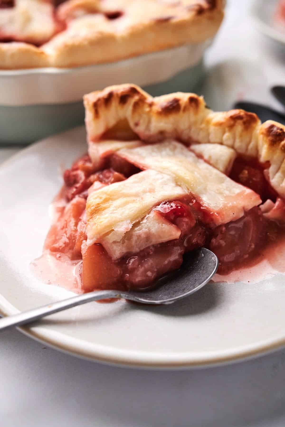 A slice of strawberry rhubarb pie with a golden-brown crust and a juicy filling served on a white plate with a spoon.