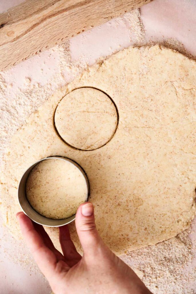 A hand holds a round cookie cutter, cutting shapes from rolled-out strawberry shortcake dough on a floured surface.